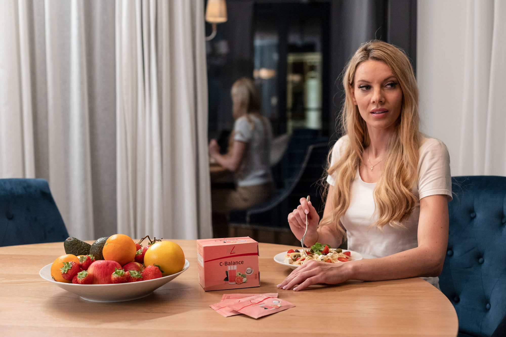 Vrouw aan tafel met FitLine C-Balance, sachets en een schaal vers fruit