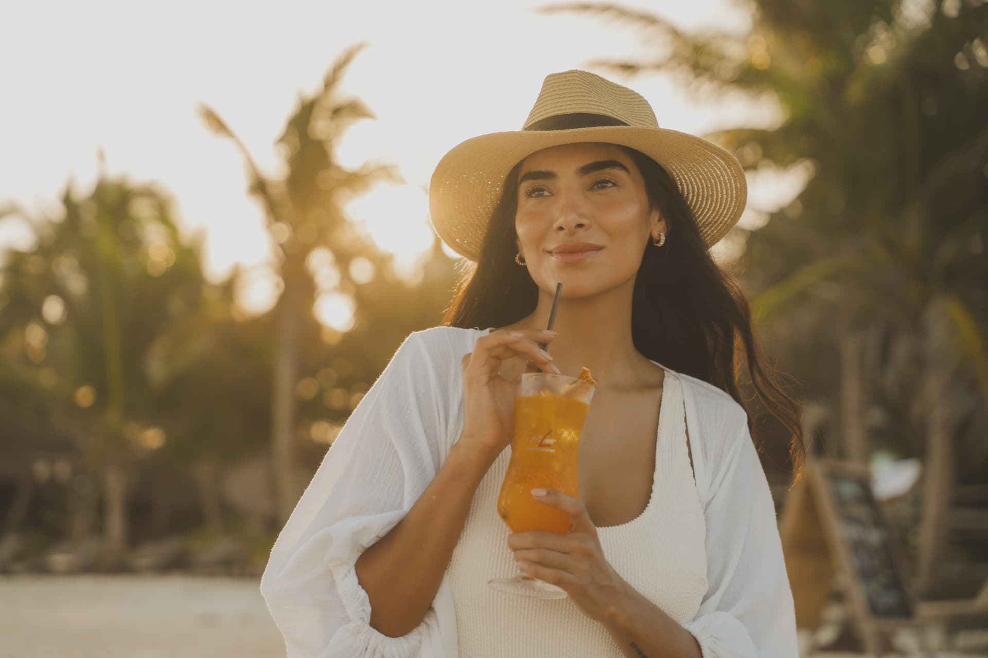 Vrouw met strohoed die een FitLine PowerCocktail drinkt in het avondlicht